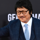 Benedict Wong waves his hand while dressed in a sharp blue suit and tie.